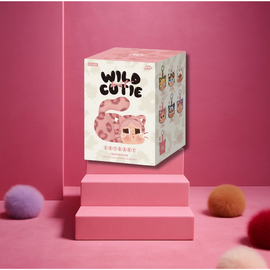 A blind box labeled "Wild Cutie Crybaby" from Pop Mart is displayed on pink steps with a pink leopard cat illustration. The background is entirely pink with colorful fluffy balls scattered on the floor.