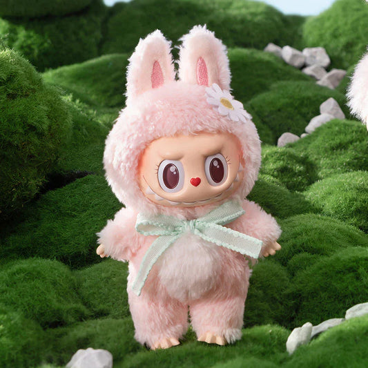 This image features an adorable Labubu figure from Pop Mart’s “The Monsters” series, dressed in a fluffy pink bunny suit with upright ears. The figure has big, expressive eyes, a red heart-shaped nose, and a mischievous smile. A mint green ribbon is tied around its neck, and a white daisy decorates one ear. The figure is posed outdoors on a lush green mossy landscape with small white stones, creating a whimsical and nature-inspired setting.