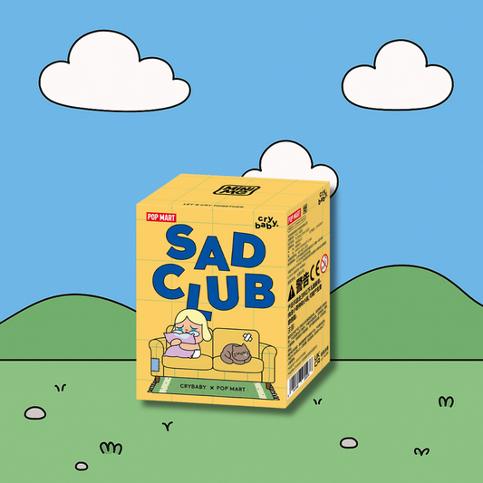 A yellow box for the "Crybaby Sad Club" series is centered on a whimsical, cartoonish outdoor background. The box's front features a character with blonde pigtails crying on a couch with a cat. The background is a blue sky with two white clouds and green hills with small, scattered outlines of plants or rocks.