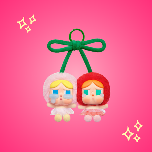 A cute double keychain featuring two plush figures with teary eyes, one in a pink outfit and the other in red, tied together with a green bow resembling cherries. Set against a bright pink background with sparkles, the design is playful and whimsical.