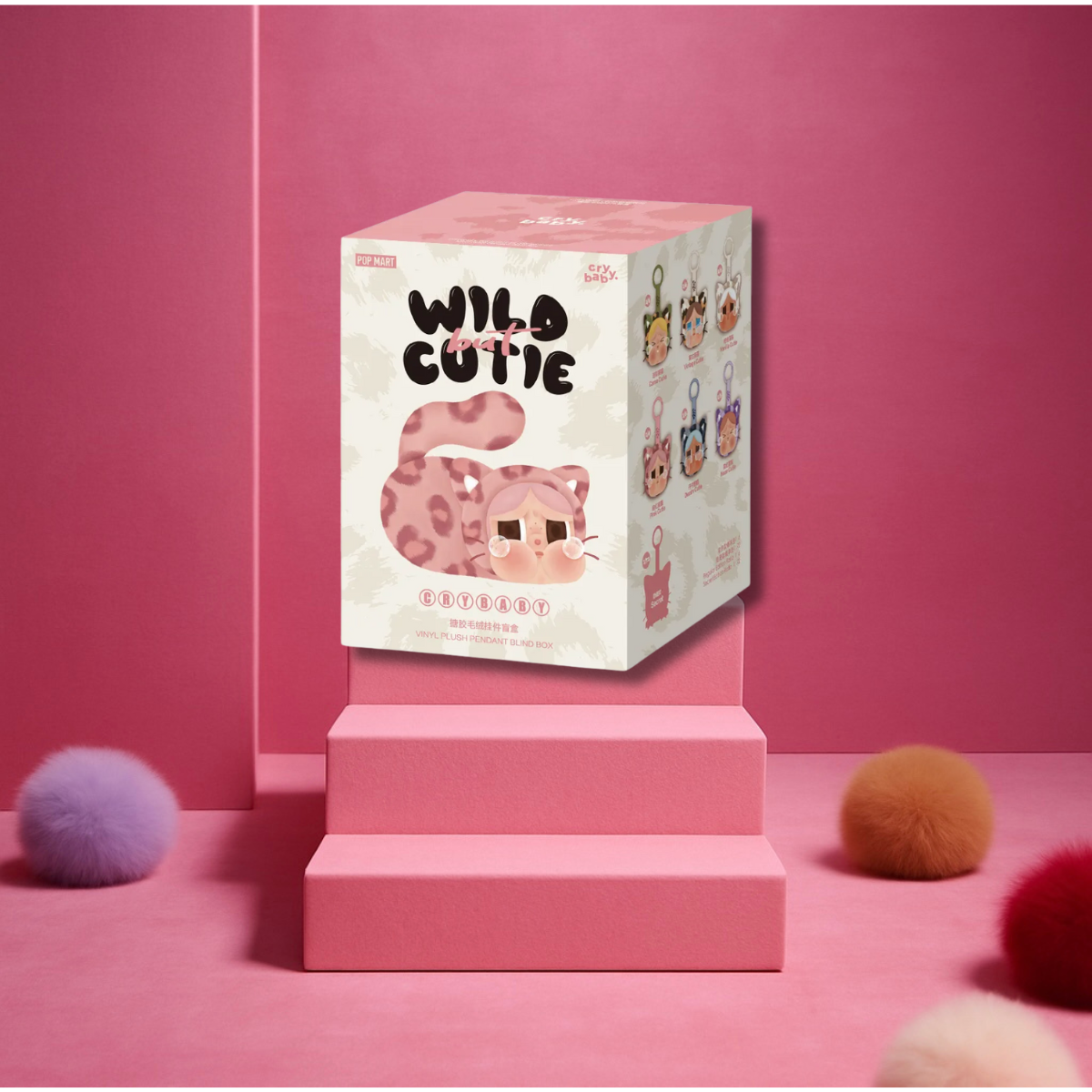 A blind box labeled "Wild Cutie Crybaby" from Pop Mart is displayed on pink steps with a pink leopard cat illustration. The background is entirely pink with colorful fluffy balls scattered on the floor.